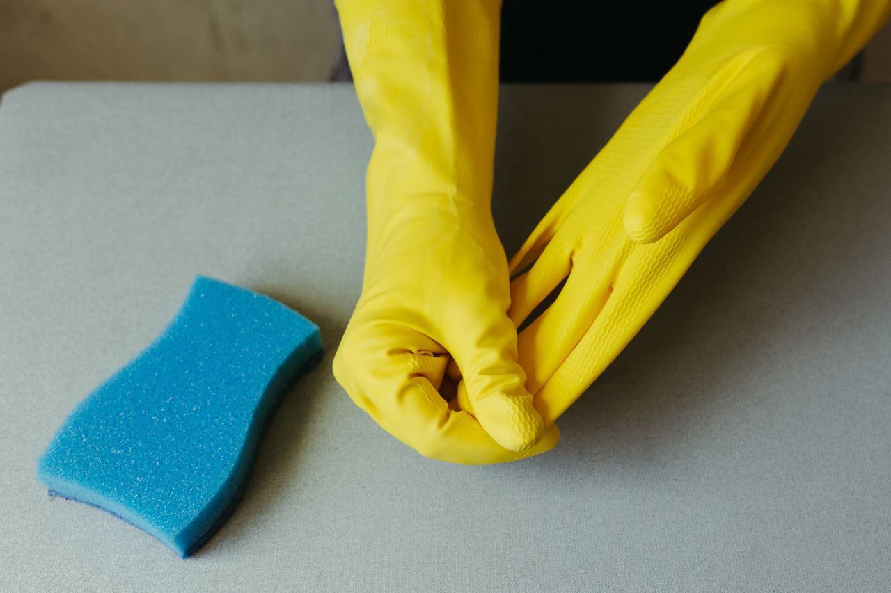 Vue rapprochée de mains portant des gants jaunes, à côté d’une éponge bleue sur une surface grise.
